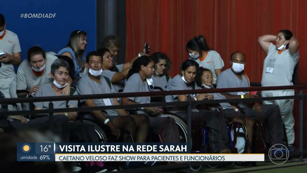 Vestindo uniformes hospitalares, pacientes em cadeiras de rodas acomodam-se em uma das galerias laterais do Teatro SARAH para assistir ao espetáculo de Caetano Veloso. Na base da imagem, vinheta mostra os dizeres: "VISITA ILUSTRE NA REDE SARAH. Caetano Veloso faz show para pacientes e funcionários."