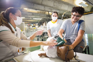 Foto tirada em enfermaria do SARAH Brasília. Sentada, profissional de saúde, usando máscara cirúrgica e vestindo uniforme, manuseia boneca de plástico, que tem um bocal de anestesia verde no rosto. Ao seu lado, menino com uniforme de paciente sorri e examina a boneca. Mais atrás, mulher com uniforme de acompanhante e máscara cirúrgica observa a cena. (Crédito: Bruno Esaki, Metrópoles).