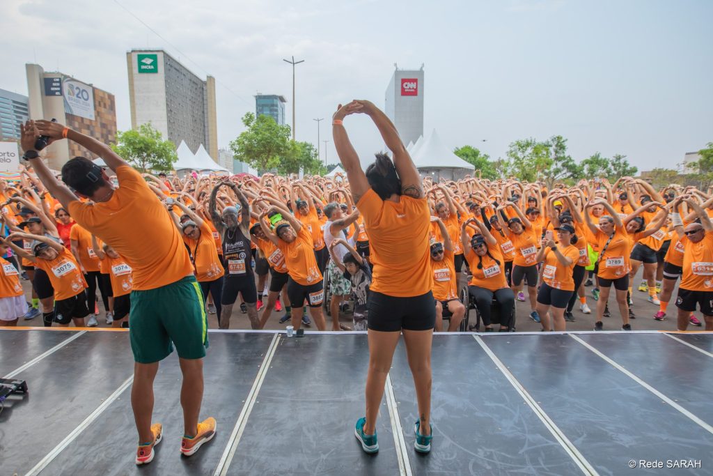 De costas, à esquerda, Caio Bonfim, e à direita, Aline Furtado, conduzindo o aquecimento com os participantes antes da corrida